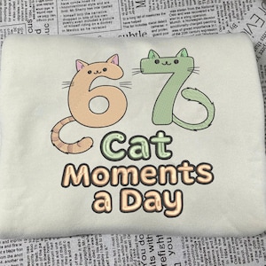 May include: Cream-colored t-shirt with the embroidered text "67 Cat Moments a Day." The numbers 6 and 7 are designed as cartoon cats, one tan and one green.
