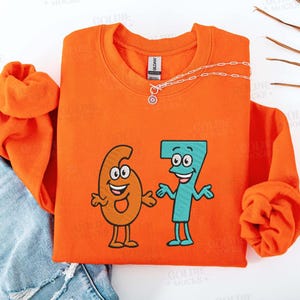 K&ouml;nnte beinhalten: Ein orangefarbenes Sweatshirt mit einer Cartoon-Grafik der Zahlen sechs und sieben, jeweils mit l&auml;chelnden Gesichtern und Armen. Eine silberne Halskette mit Sonnenanh&auml;nger befindet sich auf dem Sweatshirt. Das Sweatshirt ist mit einer Jeans gefaltet.