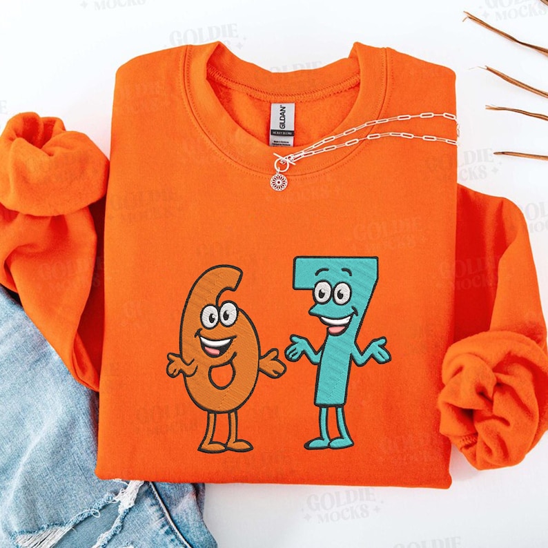 K&ouml;nnte beinhalten: Ein orangefarbenes Sweatshirt mit einer Cartoon-Grafik der Zahlen sechs und sieben, jeweils mit l&auml;chelnden Gesichtern und Armen. Eine silberne Halskette mit Sonnenanh&auml;nger befindet sich auf dem Sweatshirt. Das Sweatshirt ist mit einer Jeans gefaltet.