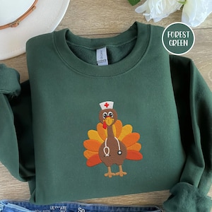 May include: Forest green sweatshirt with a turkey design wearing a nurse's hat and stethoscope. The turkey has orange, yellow, and brown feathers. The sweatshirt is folded, with the brand name visible. The words "Forest Green" are in a circle.