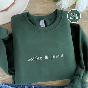 May include: Forest green sweatshirt with the words "coffee & jesus" in white script. The sweatshirt is folded on a wooden surface. A white hat and flower are in the background.