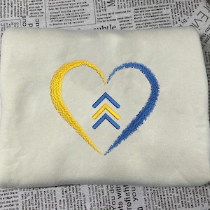 May include: Cream-colored sweatshirt featuring an embroidered heart design. The heart is split into yellow and blue sections, with three upward-pointing arrows in the center. The design is centered on the fabric.