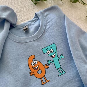 K&ouml;nnte beinhalten: Hellblaues Sweatshirt mit den Cartoon-Zahlen sechs und sieben. Die Zahl sechs ist orange und die Zahl sieben ist t&uuml;rkis. Das Sweatshirt hat einen Rundhalsausschnitt und lange &Auml;rmel.