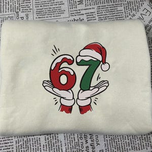 May include: Cream-colored sweatshirt featuring the number "67" in a festive design. The "6" is red, and the "7" is green, both held by cartoon hands with white gloves and red cuffs. A Santa hat tops the "7".