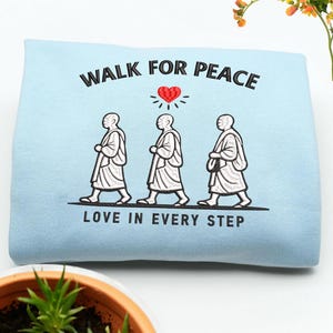 May include: A pale blue sweatshirt featuring the embroidered words "WALK FOR PEACE" above three walking figures and a heart. Below the figures, the text reads "LOVE IN EVERY STEP". The figures are in white.