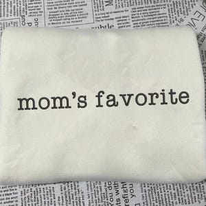 May include: Cream-colored t-shirt with the embroidered text "mom's favorite" in a dark brown color. The shirt is made of a soft material and is suitable for casual wear.