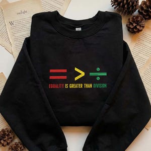 May include: Black sweatshirt with the embroidered text "EQUALITY IS GREATER THAN DIVISION." The design features an equal sign in red, a greater than symbol in yellow, and a division symbol in green.