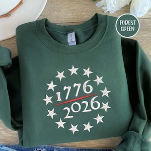 May include: Forest green sweatshirt with a crew neck. The front features a white embroidered design of stars encircling the numbers "1776" and "2026" with a red line through the middle. The sweatshirt is folded on a wooden surface.