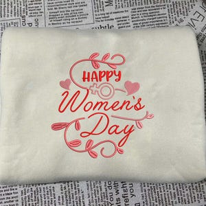 Puede incluir: Sudadera crema con texto bordado en rojo "HAPPY Women's Day", con un símbolo femenino y corazones. Un diseño festivo para celebrar a las mujeres.