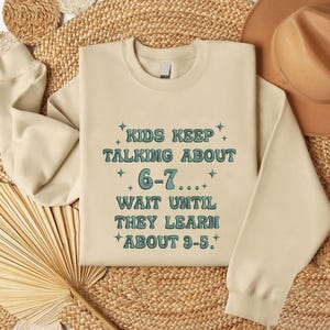 May include: A cream-colored sweatshirt with the text "KIDS KEEP TALKING ABOUT 6-7... WAIT UNTIL THEY LEARN ABOUT 9-5." in teal lettering. The sweatshirt is displayed on a woven surface, with a hat visible in the background.