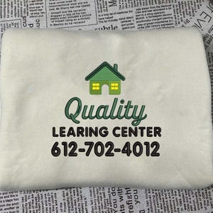 May include: Cream-colored rectangular item with the words "Quality LEARNING CENTER" in black and green, along with a house graphic and a phone number. The item appears to be a pillow or a similar fabric product.