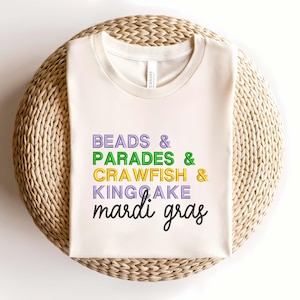 Puede incluir: Camiseta color crema con las palabras "BEADS & PARADES & CRAWFISH & KING CAKE mardi gras" en letras moradas, verdes, amarillas y negras. La camiseta está doblada sobre una superficie circular tejida.