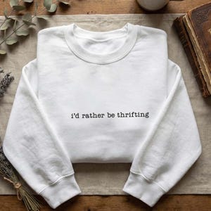 Puede incluir: Sudadera blanca de cuello redondo con la frase "i'd rather be thrifting" impresa en negro. La sudadera es de material suave y tiene mangas largas. La imagen está tomada desde arriba.