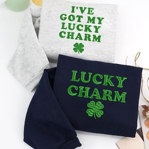 May include: Two sweatshirts are shown, one light grey and one navy blue. The light grey sweatshirt has the text "I'VE GOT MY LUCKY CHARM" embroidered in green, with a green shamrock. The navy blue sweatshirt says "LUCKY CHARM" with a green shamrock.