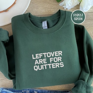 Puede incluir: Sudadera verde bosque con el texto blanco "LEFTOVER ARE FOR QUITTERS". La sudadera doblada parece estar hecha de un material suave. Un sombrero blanco y una flor están en el fondo.