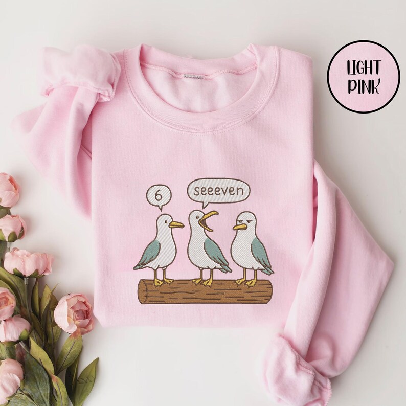 May include: Light pink sweatshirt featuring a cartoon design of three seagulls on a log. One seagull has a thought bubble with the number 6, while another says "seeeven". The sweatshirt has long sleeves and a round neckline.