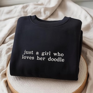 May include: Black sweatshirt with the white text "just a girl who loves her doodle". The crew neck sweatshirt is folded and resting on a light-colored surface. The text is in a simple, sans-serif font.