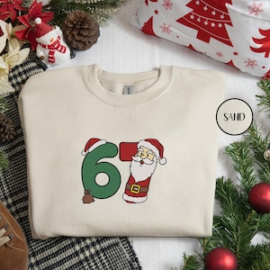 May include: A sand-coloured sweatshirt with a festive Christmas design. The design features the numbers "67" with the "6" in green and the "7" designed as Santa Claus. The Santa Claus illustration includes a red hat, white beard, and a brown belt.