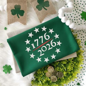 May include: A green fabric item with the numbers "1776" and "2026" in white, encircled by white stars, and a red line. The item is on a white surface with shamrock decorations and gold coins.