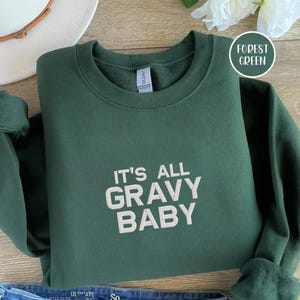 Puede incluir: Sudadera verde bosque con el texto blanco "IT'S ALL GRAVY BABY". La sudadera está doblada. La imagen también muestra un sombrero blanco y una flor en el fondo. La sudadera parece estar hecha de un material suave.
