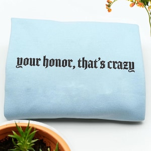 May include: A light blue sweatshirt with the phrase "your honor, that's crazy" embroidered in black gothic lettering. The sweatshirt is folded and appears soft, with a simple, clean design.