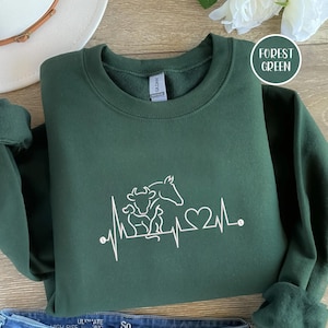 Puede incluir: Sudadera verde bosque con un gráfico blanco de una vaca, un caballo, un perro y una línea de latidos. El texto "Forest Green" está en un círculo blanco. La sudadera está doblada sobre una superficie de madera.