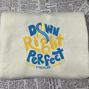 May include: Cream-colored sweatshirt with the embroidered phrase "Down Right Perfect" in blue and yellow, with a blue and yellow ribbon design. The text "#DOWN SYNDROME AWARENESS" is also embroidered.