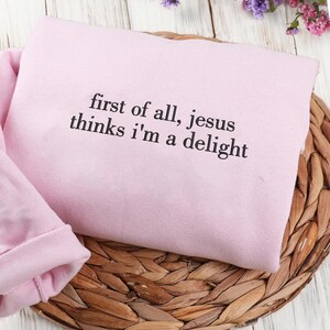 Pu&ograve; includere: Una felpa rosa chiaro con la scritta nera "first of all, jesus thinks i'm a delight". Il capo piegato &egrave; appoggiato su un sottobicchiere rotondo e intrecciato, con fiori sullo sfondo.