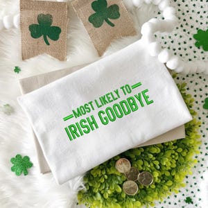 May include: A white St. Patrick's Day themed kitchen towel with the text "MOST LIKELY TO - IRISH GOODBYE" in green. The towel is on top of a beige towel, with shamrock decorations and gold coins with shamrocks.