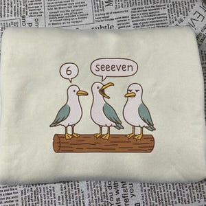 May include: Embroidered sweatshirt featuring three cartoon seagulls perched on a log. One seagull has a thought bubble with the number "6", and another says "seeeven". The seagulls are white, gray, and teal.