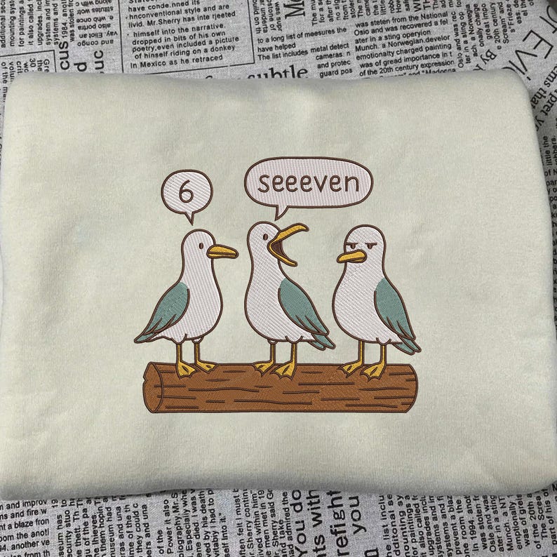 May include: Embroidered sweatshirt featuring three cartoon seagulls perched on a log. One seagull has a thought bubble with the number "6", and another says "seeeven". The seagulls are white, gray, and teal.
