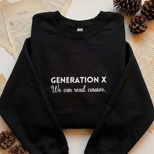 May include: Black sweatshirt with the text "GENERATION X We can read cursive." in white. The sweatshirt is displayed on a surface with open books and pinecones.