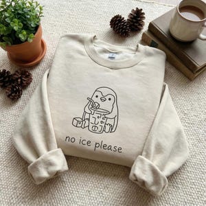 Op de afbeelding: Crèmekleurige sweatshirt met een zwarte afbeelding van een pinguïn die uit een beker met een rietje drinkt. De tekst "no ice please" staat onder de pinguïn. De sweatshirt heeft een ronde hals en lange mouwen met opgerolde manchetten. De achtergrond bevat een plant, dennenappels en een mok.
