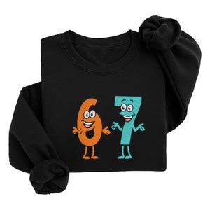 K&ouml;nnte beinhalten: Schwarzes Sweatshirt mit den Zahlen 6 und 7 als Zeichentrickfiguren. Die Zahl 6 ist orange mit einem l&auml;chelnden Gesicht und Beinen. Die Zahl 7 ist t&uuml;rkis mit einem l&auml;chelnden Gesicht, Armen und Beinen. Das Sweatshirt hat einen Rundhalsausschnitt.