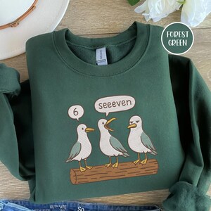 May include: Forest green sweatshirt with a graphic of three seagulls on a log. One seagull has a speech bubble that says "seeeven", and another has a speech bubble with the number "6".
