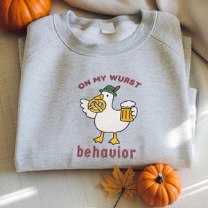 Può includere: Una felpa grigio chiaro con la scritta "ON MY WURST behavior" in lettere color marrone. La felpa presenta un'anatra dei cartoni animati con un cappello e che tiene un pretzel e una birra. Due zucche e una foglia sono sullo sfondo.