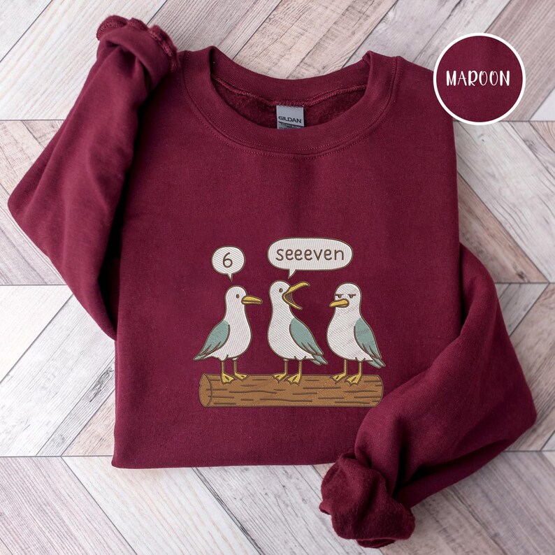 May include: Maroon sweatshirt with a graphic of three seagulls on a log. One seagull has its beak open, and speech bubbles say "6" and "seeeven". The sweatshirt is a solid maroon color.