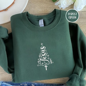 May include: Forest green sweatshirt with a white Christmas tree design that spells out "Jesus". The design includes stars, snowflakes, and a cross. The sweatshirt is folded on a wooden surface.