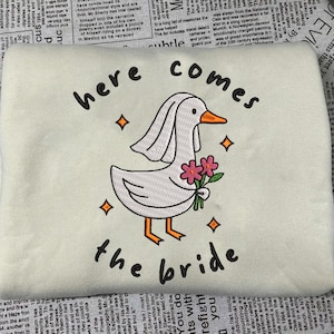 Pode incluir: Camisola creme com um desenho bordado com um ganso usando um véu e segurando flores, com o texto "here comes the bride". O design inclui detalhes em laranja e rosa.