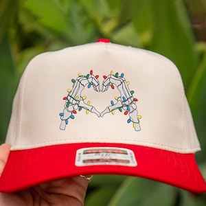 May include: A beige baseball cap with a red brim and a red button on top. The cap features a graphic of two skeleton hands forming a heart shape, decorated with colorful Christmas lights.