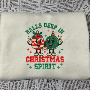May include: Cream-colored sweatshirt with an embroidered design featuring two cartoon Christmas ornaments with arms and legs. The text reads "BALLS DEEP IN CHRISTMAS SPIRIT" in red and green.