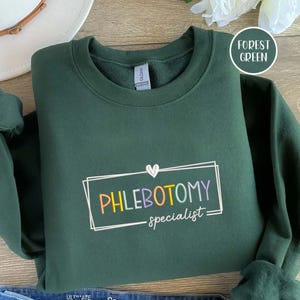 Könnte beinhalten: Ein dunkelgrünes Sweatshirt mit dem Text "PHLEBOTOMY specialist" in einem rechteckigen Design. Der Text ist mehrfarbig, mit Gelb, Orange, Lila und Grün. Ein weißes Herz befindet sich über dem Wort "PHLEBOTOMY".