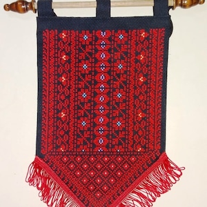 May include: A red and black embroidered wall hanging with a fringe border. The hanging is attached to a wooden dowel rod with black fabric loops.