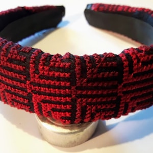 Può includere: Una fascia per capelli a fantasia rossa e nera. La fascia presenta un motivo geometrico ripetuto in filo rosso su sfondo nero. La fascia stessa è nera e sembra essere realizzata in un materiale robusto.