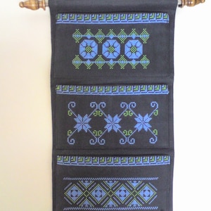 May include: Black wall hanging with three pockets, each with a different embroidered design in blue and green thread. The pockets are framed with a decorative border. The hanging has a wooden dowel rod at the top and a fringe at the bottom.