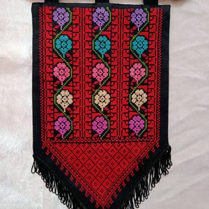 May include: A red and black embroidered wall hanging with a floral pattern. The design features colorful flowers in purple, teal, pink, and cream. The hanging has a black fringe at the bottom and is suspended from a wooden rod.