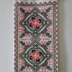 May include: A white fabric wall hanging with a wooden dowel rod at the top. The fabric features a repeating pattern of pink, green, and yellow flowers with geometric designs.