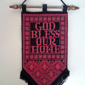 May include: A black and red embroidered wall hanging with the text "GOD BLESS OUR HOME" in pink and green lettering. The hanging has a wooden dowel rod and fringe.