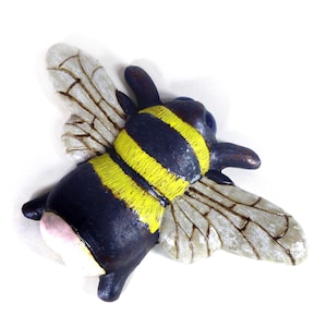 May include: A ceramic bee ornament with a black body, yellow stripes, and translucent wings. The bee has a pink abdomen and is set against a white background. The wings have detailed vein patterns.