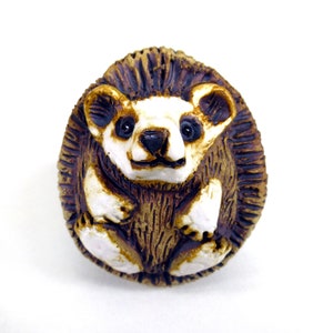 May include: A ceramic hedgehog figurine painted in brown and white. The hedgehog is curled up with its head and feet visible.
